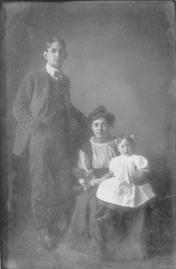 Svartvitt foto. En ung man står upp bakom en ung kvinna som sitter med ett litet barn i famnen