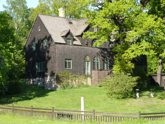 Ett stort mörkt brunt trevåningshus med tre stora kyrkolika fönster på framsidan. Solen lyser, stora ekar skuggar fasaden